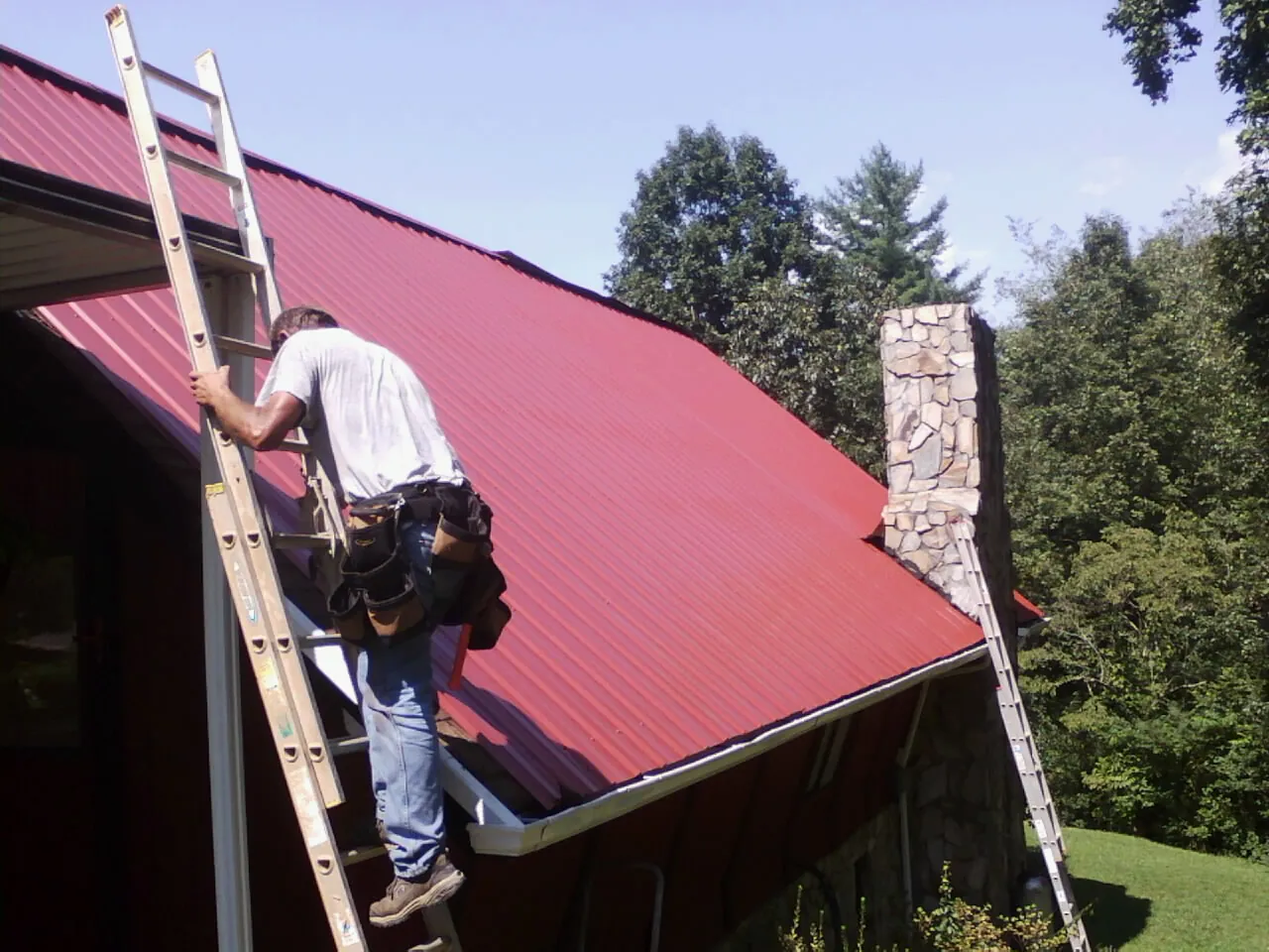 Roofer on a ladder beside a red metal roof and stone chimney — Soffit & Fascia Repair by Roofing Brothers in Manahawkin